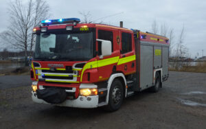 Kuvassa paloauto hiekkamaalla hälytysvalot päällä. Taustalla meri ja pilvinen taivas